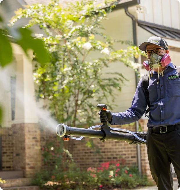 A worker in protective gear sprays pest control in a garden outside a house on a sunny day.