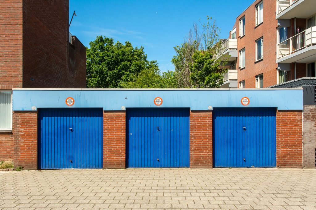 Three blue garage doors with brick walls below an apartment building, with no parking signs above each door.